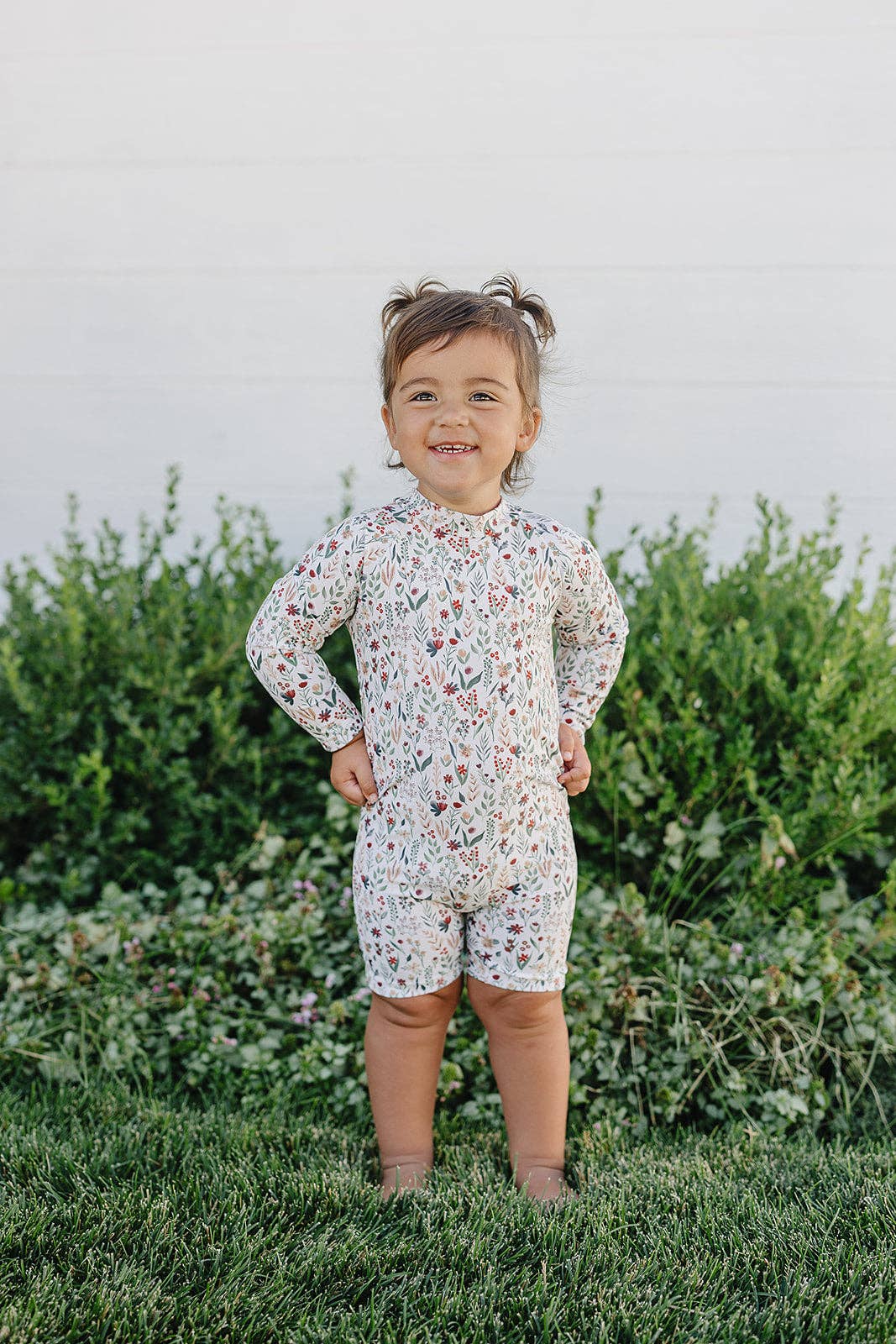Child wearing a floral romper standing outdoors with greenery in the background