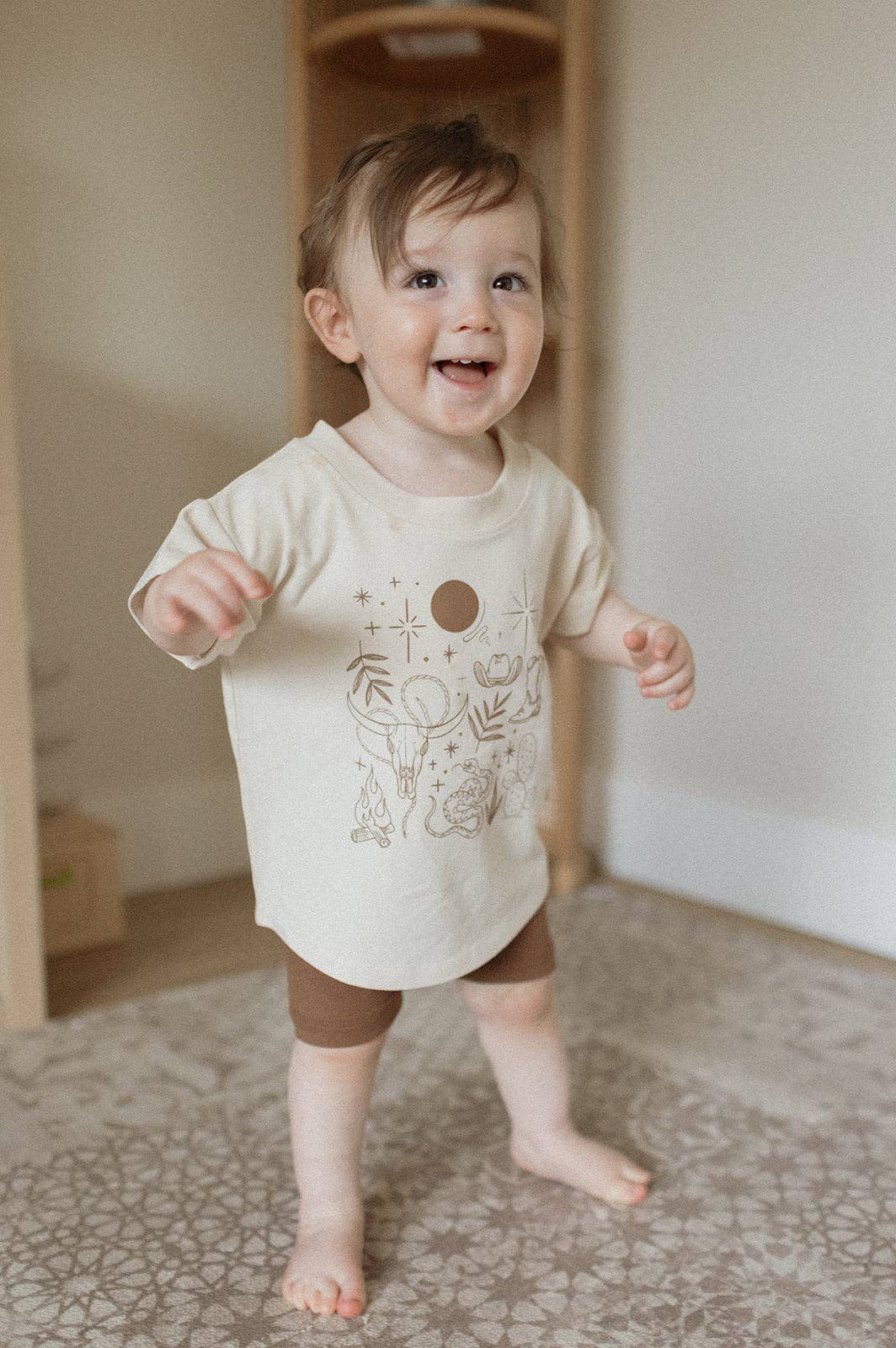 
                  
                    Baby wearing a white t-shirt with a design, standing on a patterned floor.
                  
                