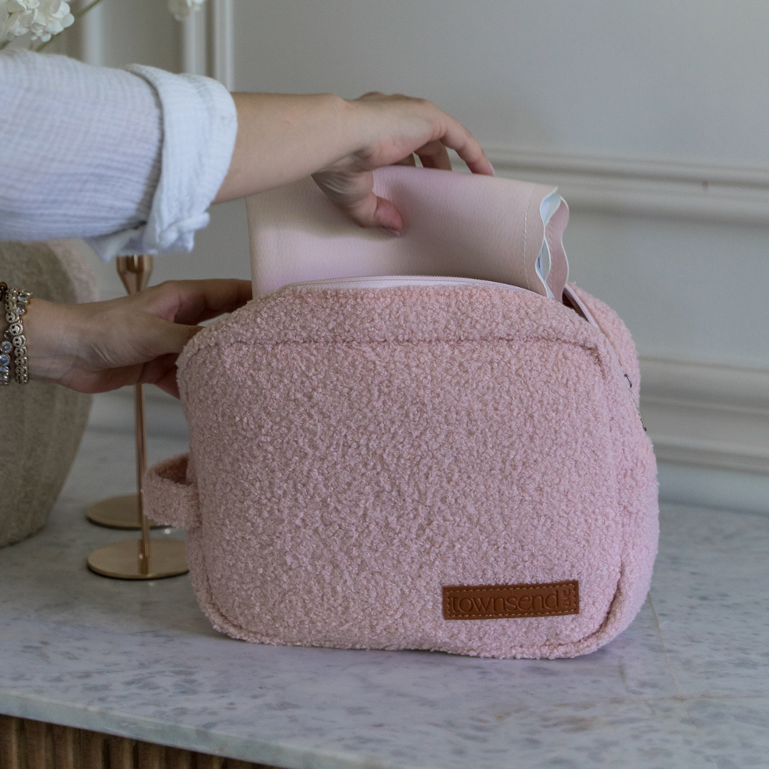 
                  
                    Pink teddy bag with a brown label on a marble surface, held by a person.
                  
                