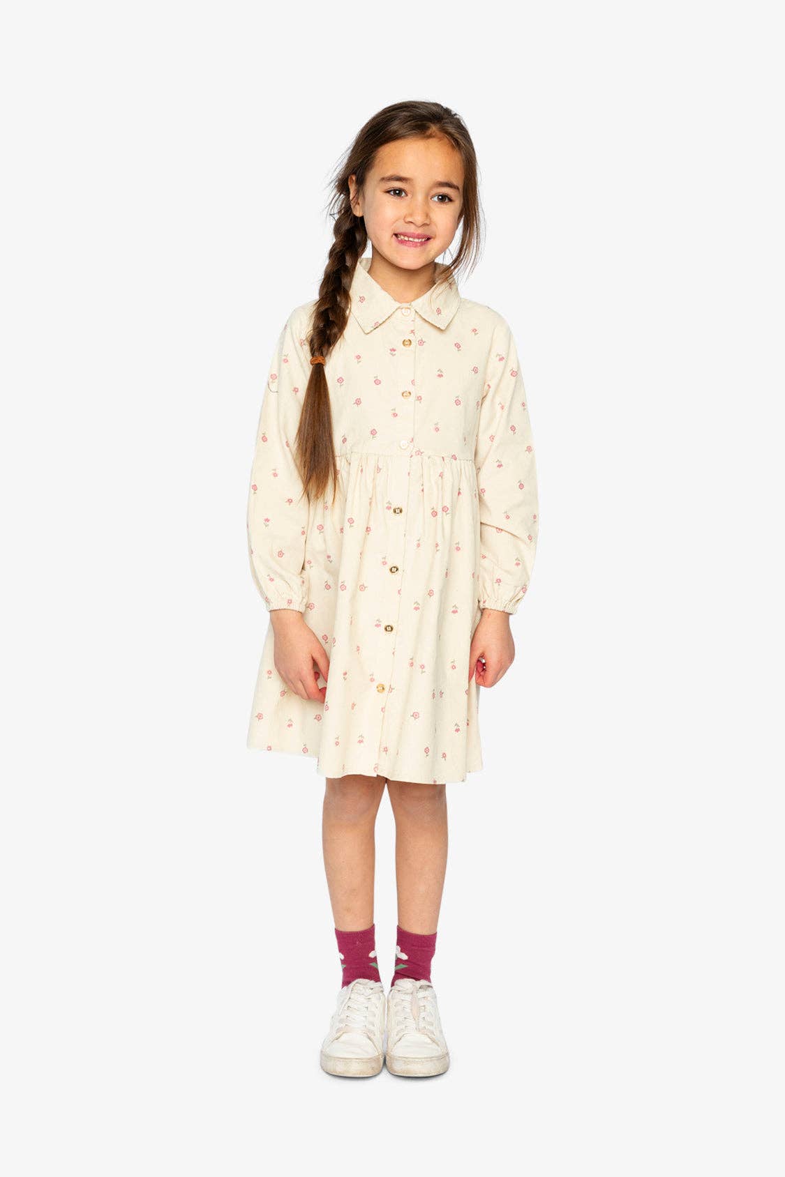 
                  
                    Young girl wearing a cream dress with a pattern, standing against a white background.
                  
                