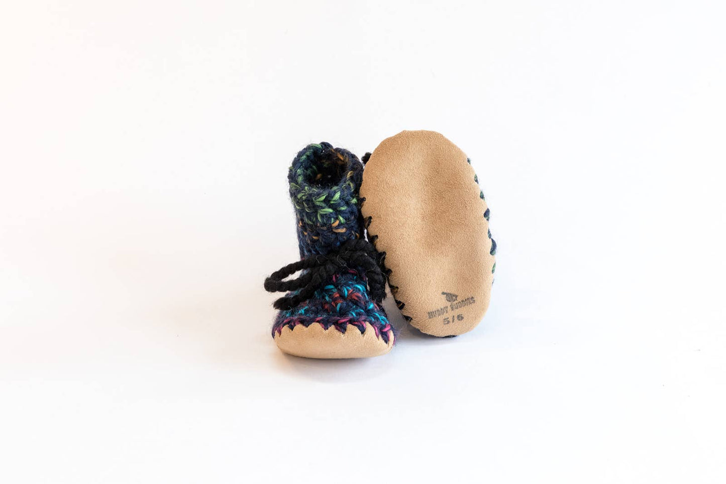 
                  
                    A pair of handmade baby booties on a white background. One slipper is shown upright, displaying its dark blue and purple patterned leather upper with black laces, while the other lies on its tan suede sole.
                  
                
