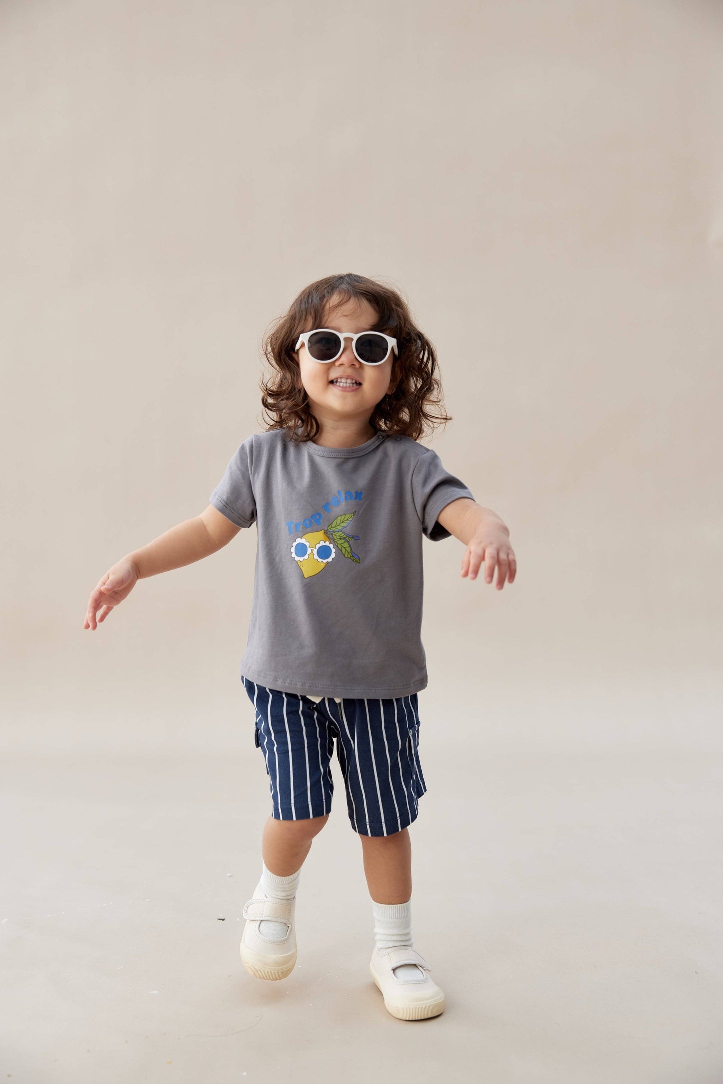 
                  
                    Child wearing a gray t-shirt with a colorful design and striped shorts on a beige background.
                  
                