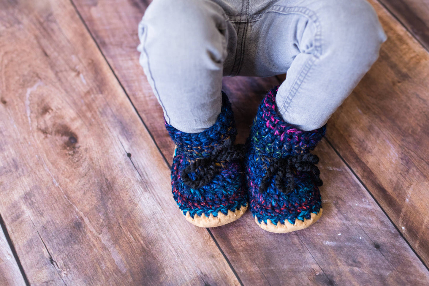 
                  
                    A close-up of a child's legs in light gray jeans, wearing handmade, multicolored crocheted booties with beige soles, standing on a rustic wooden floor.
                  
                