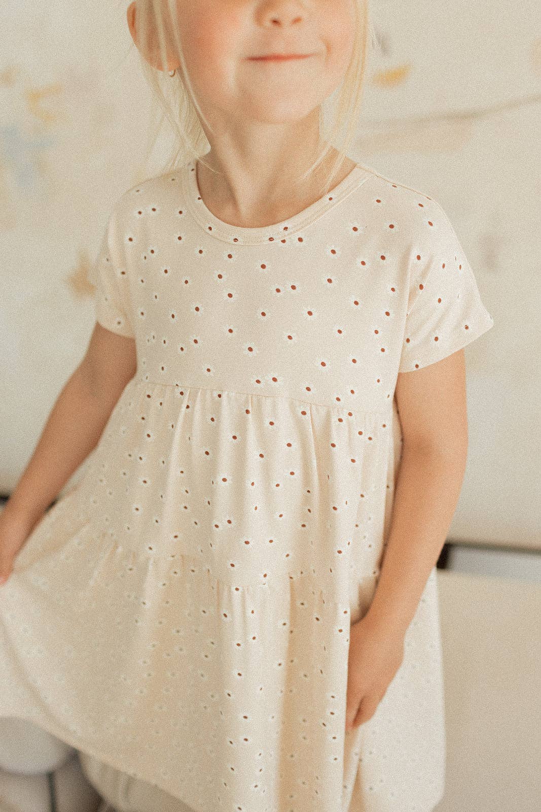 
                  
                    Young girl wearing a white dress with small patterns, standing against a neutral background.
                  
                