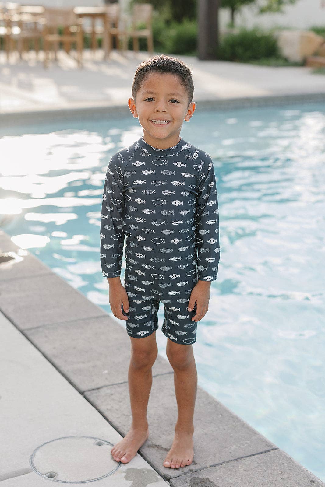Child wearing a navy blue swimsuit with fish pattern standing by a pool.