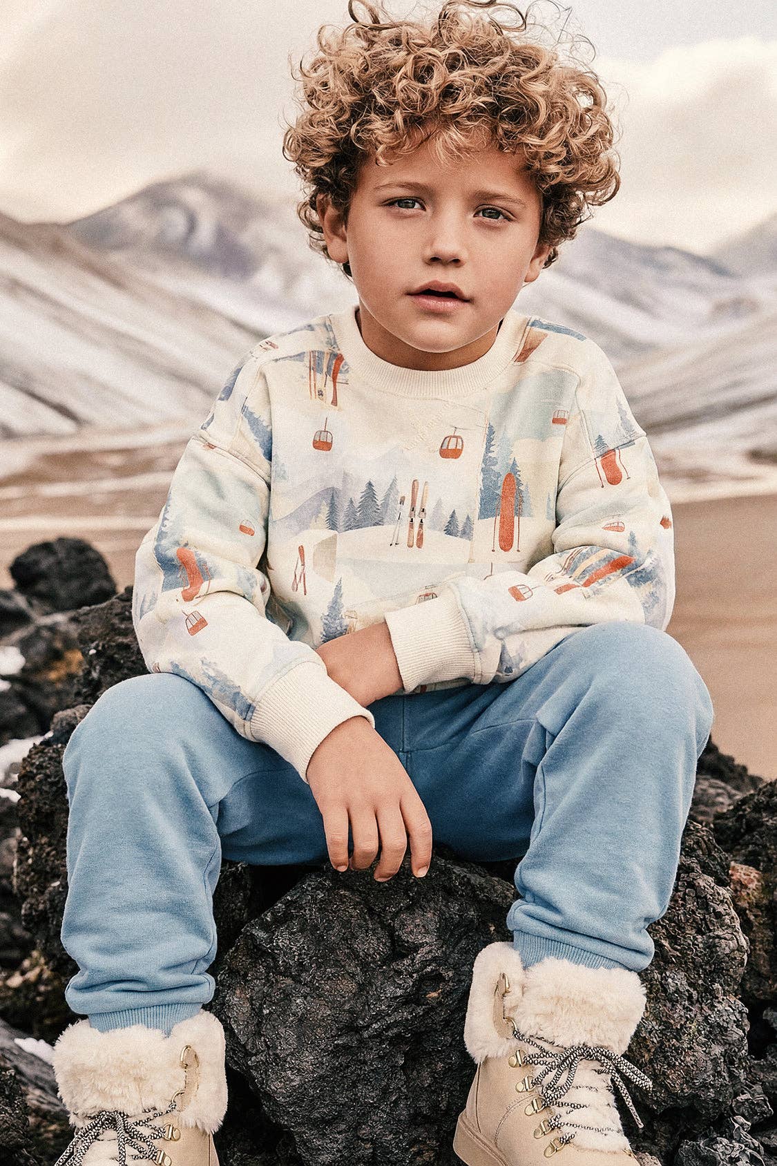 
                  
                    A young boy with curly light brown hair sits on a dark rock in a mountainous outdoor setting, wearing a cream-colored ski-themed sweatshirt with orange and blue details, light blue sweatpants, and beige boots with white faux-fur trim.
                  
                