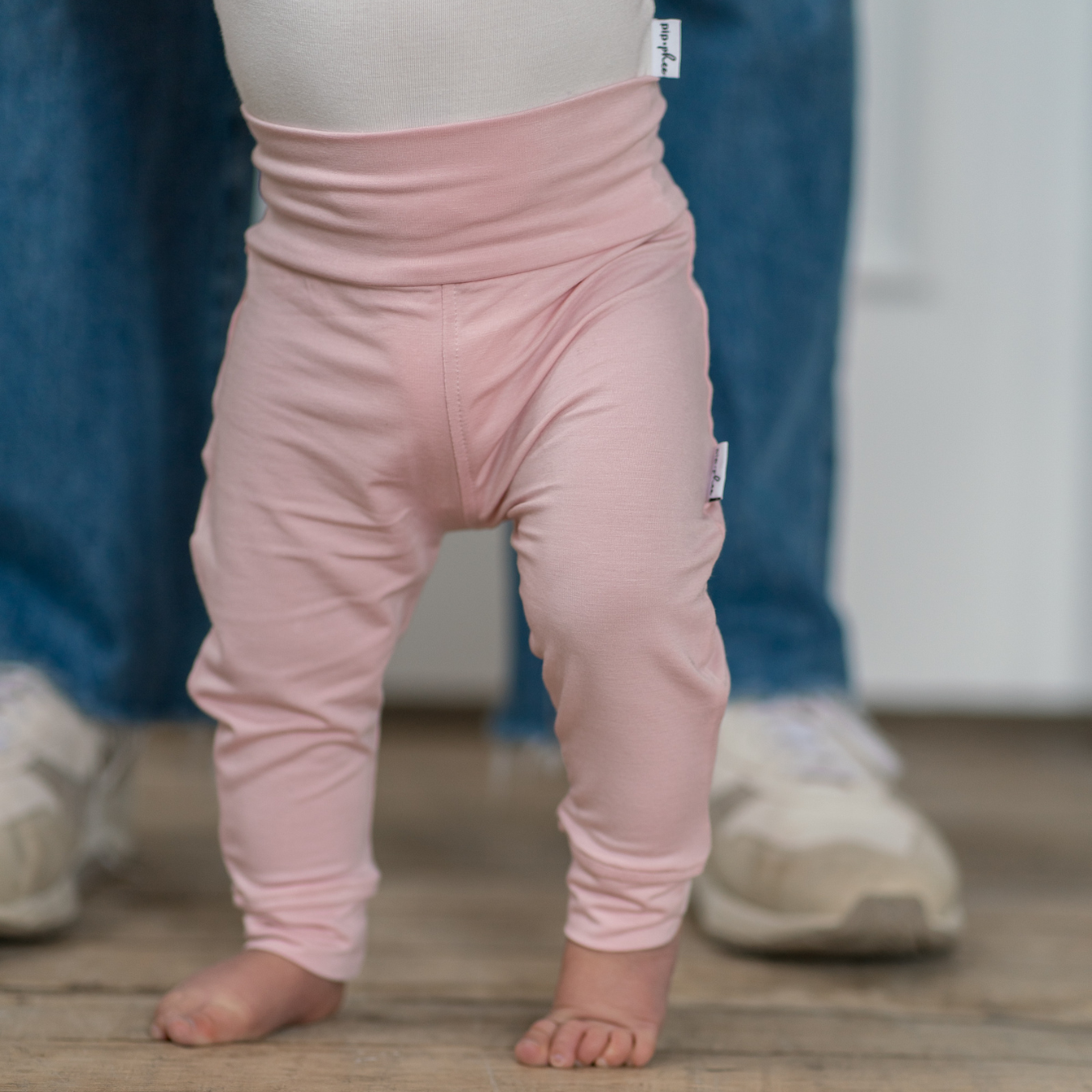 
                  
                    Child wearing pink pants standing on a wooden floor with a blurred background.
                  
                