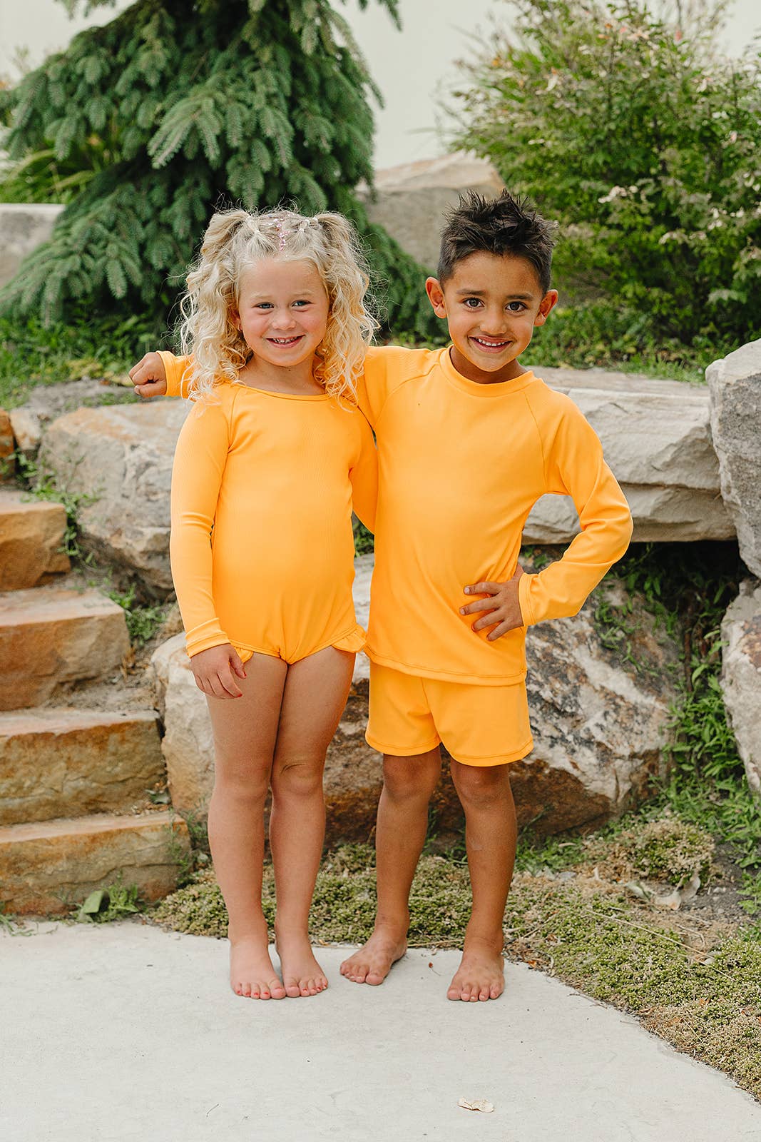 
                  
                    Two children in matching orange swim outfits standing outdoors on a stone path with greenery in the background.
                  
                