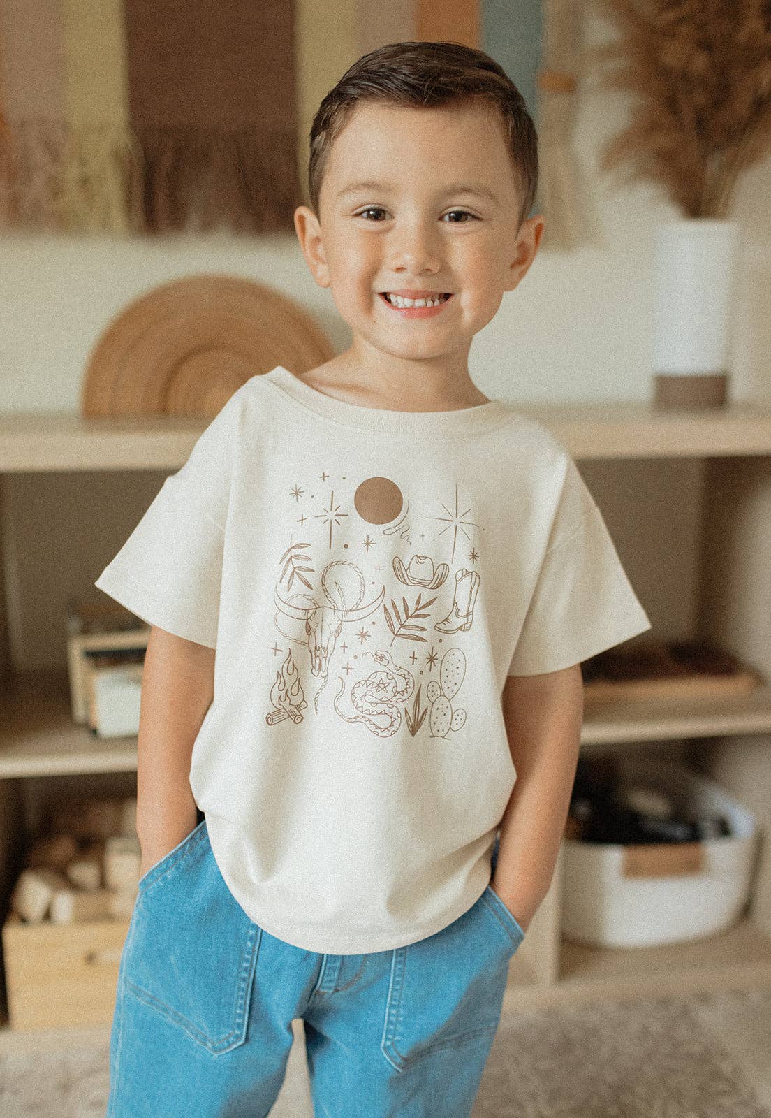 
                  
                    Child wearing a white t-shirt with a design, standing in a room with wooden shelves and decor.
                  
                