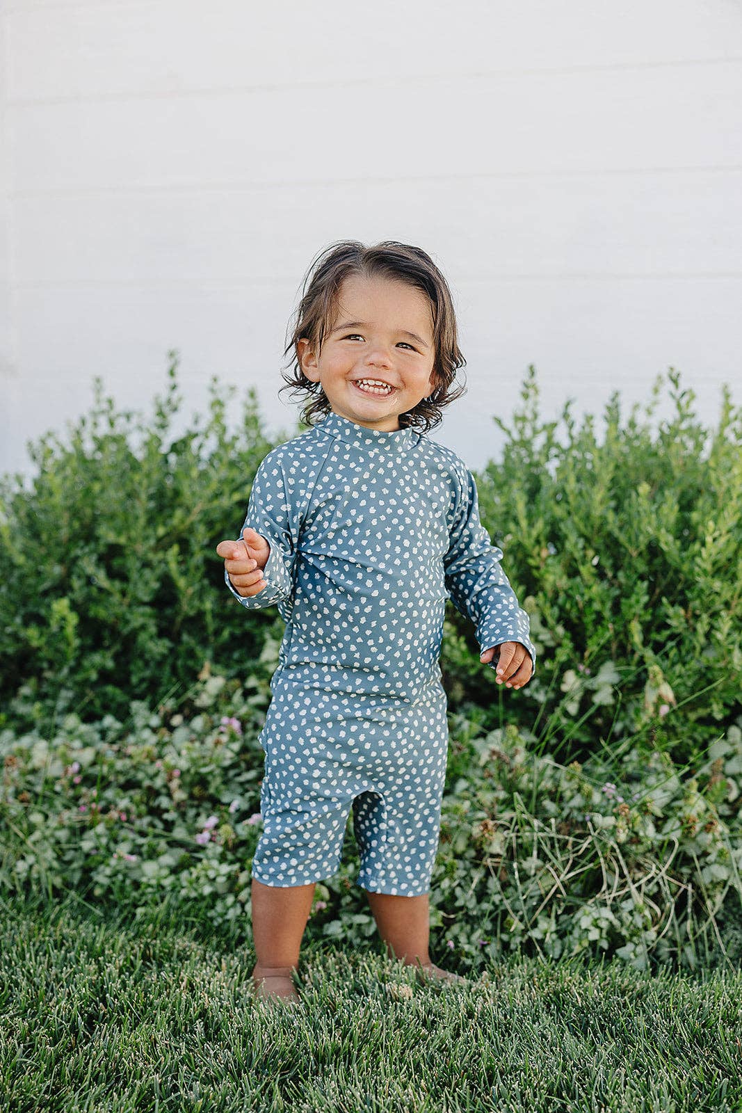 Child wearing a blue polka dot sunsuit standing in front of green bushes.
