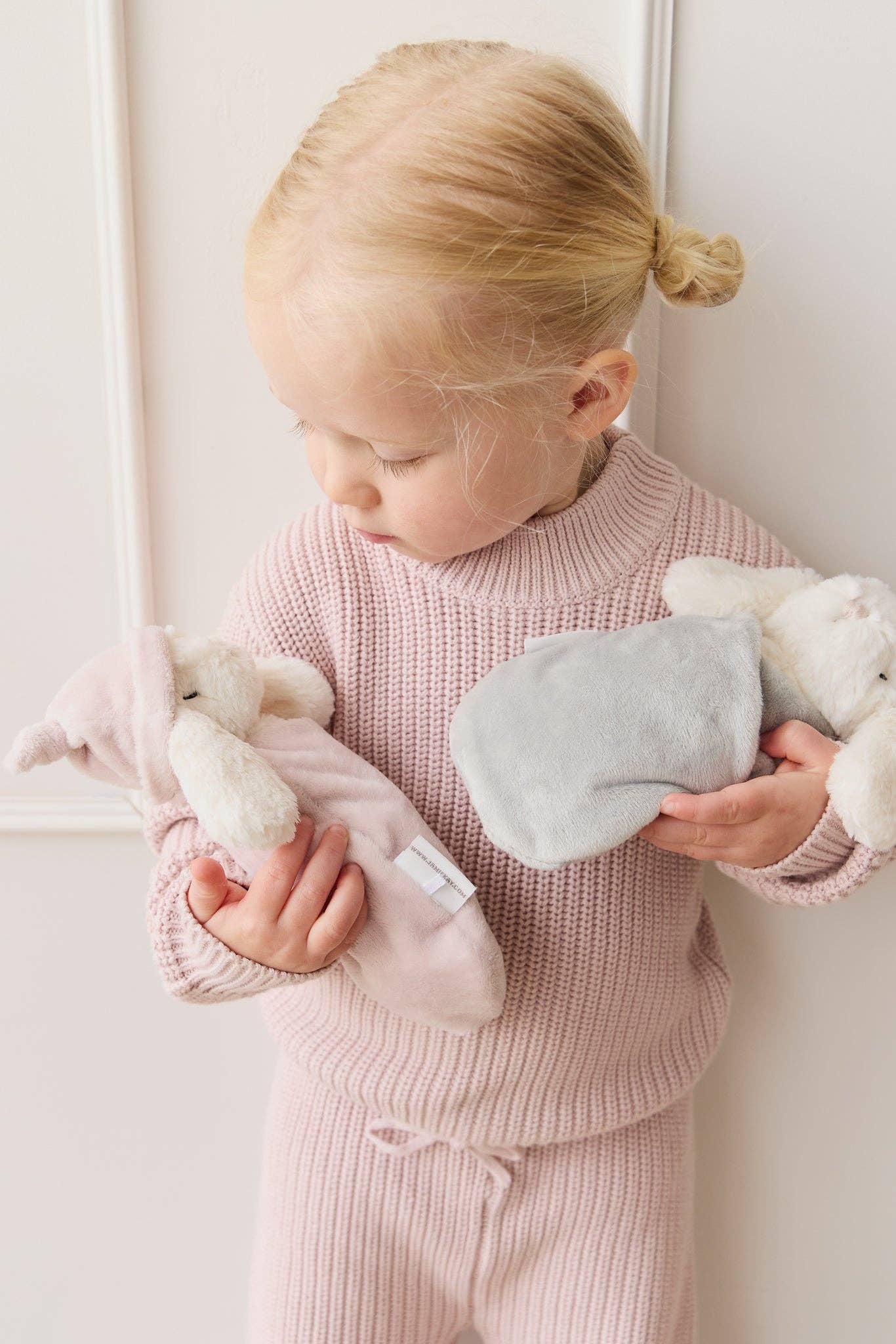 
                  
                    Child in pink outfit holding plush toys against a white wall.
                  
                