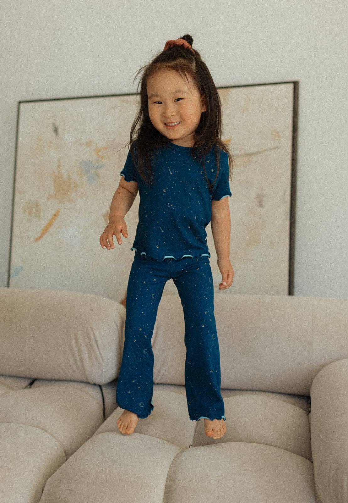 
                  
                    Child wearing a blue outfit standing on a beige sofa with a neutral wall and abstract art in the background.
                  
                