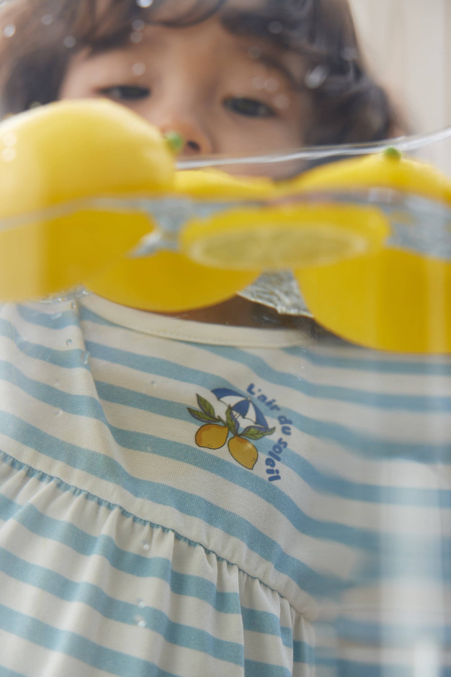 
                  
                    Child holding lemons behind a glass container with a striped dress featuring lemon designs.
                  
                