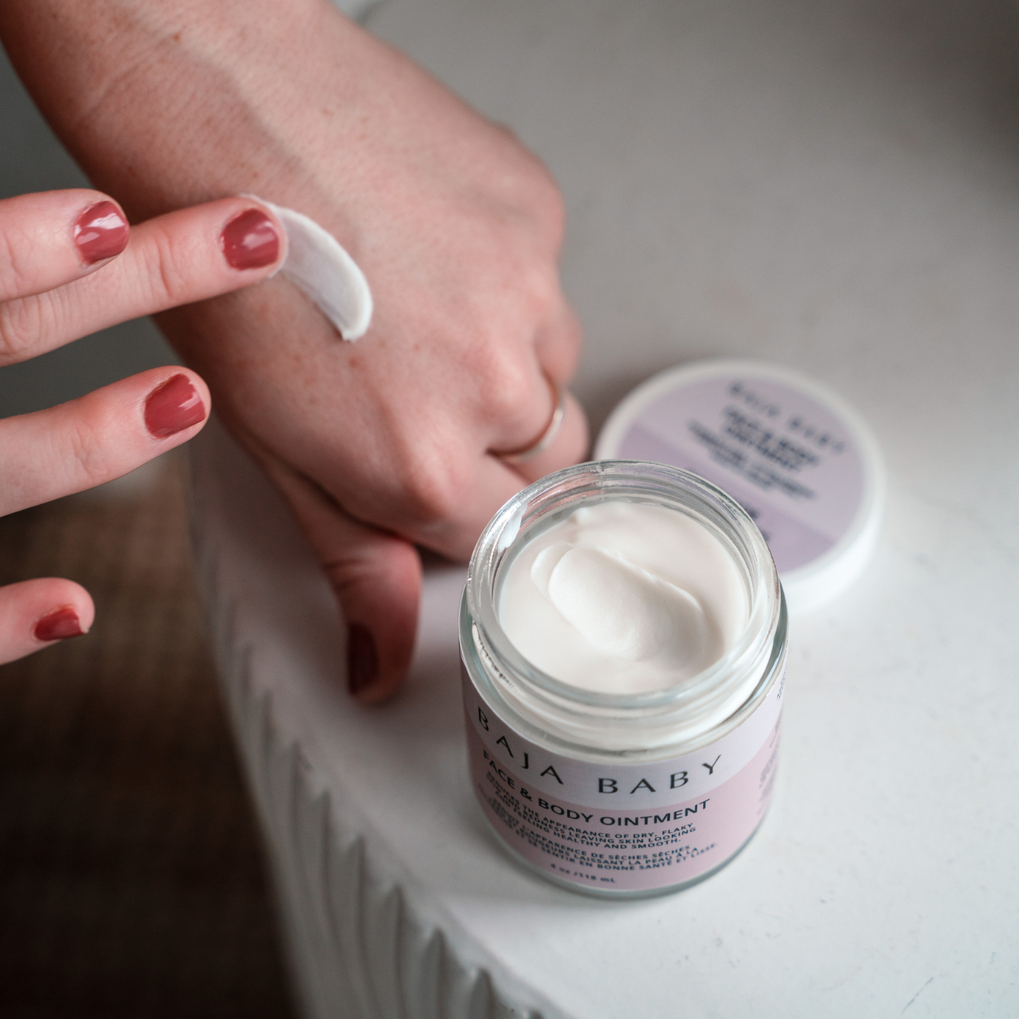 
                  
                    Person applying cream from a jar labeled 'Baja Baby' on a neutral background.
                  
                