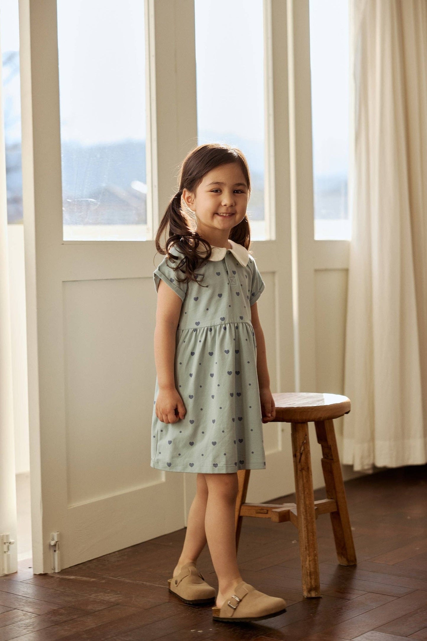 
                  
                    Young girl in a light blue dress standing in a room with large windows and a wooden stool.
                  
                