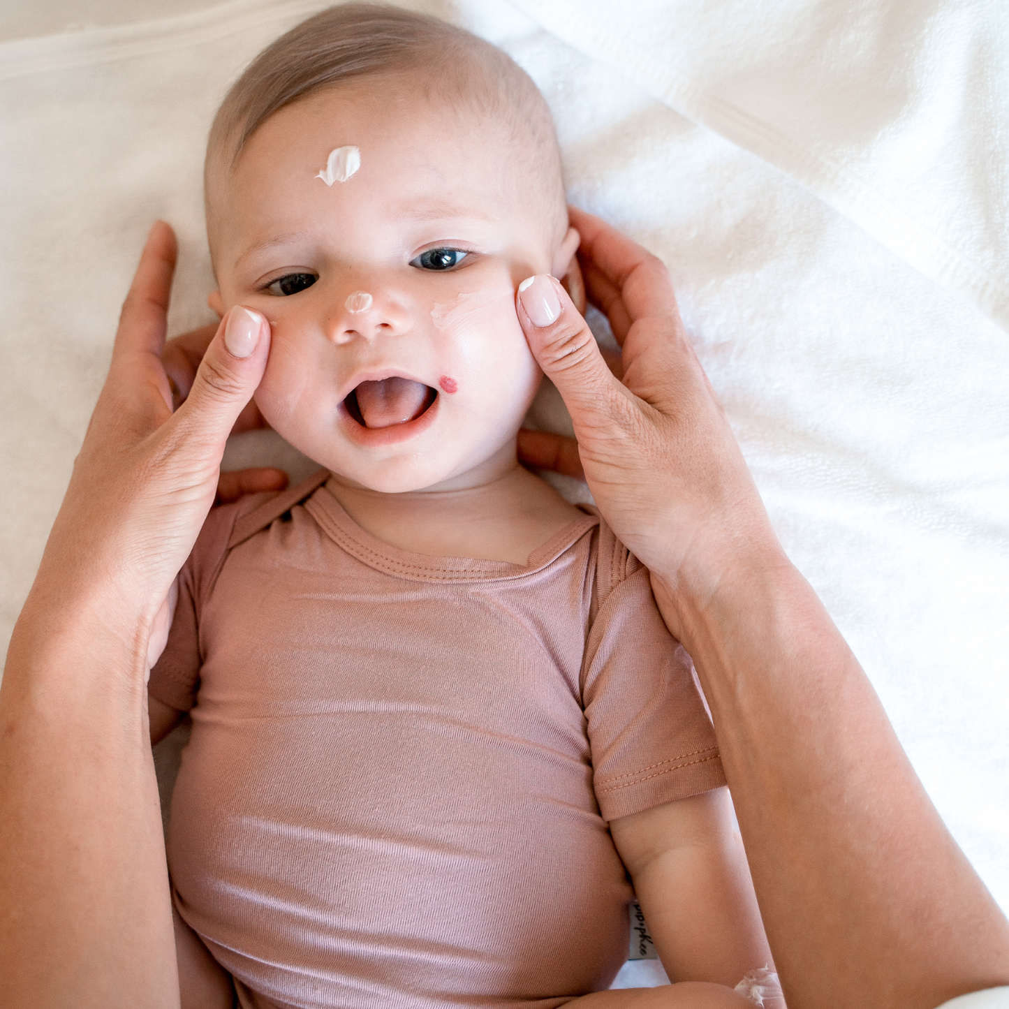 
                  
                    Baby in a pink onesie with hands gently touching its face on a soft surface.
                  
                