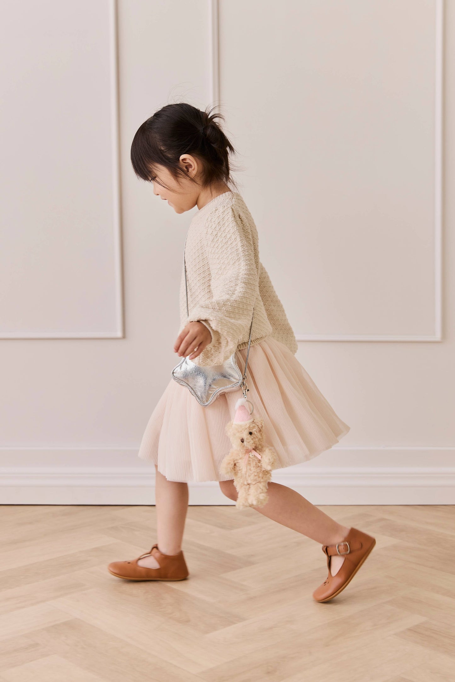 
                  
                    Child in a beige dress with a teddy bear and small bag on a wooden floor.
                  
                