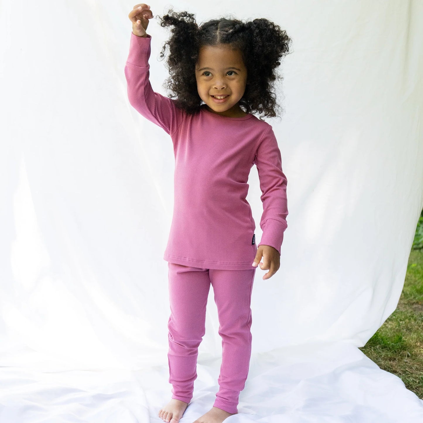 
                  
                    A young girl with curly dark hair stands barefoot in front of a white fabric backdrop, wearing a matching pink long-sleeve top and pants set.
                  
                
