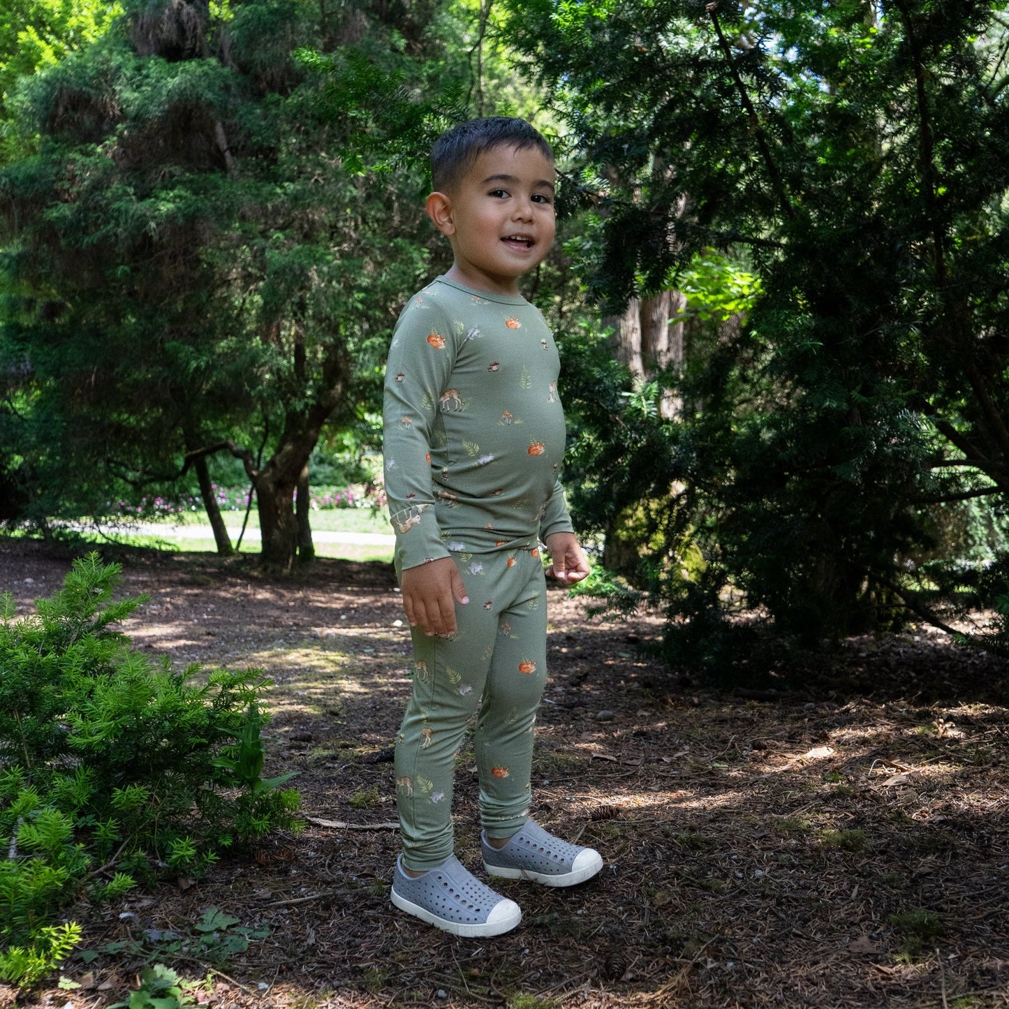 
                  
                    A young child wearing a green long-sleeve pajama set with a small animal print, standing in a wooded outdoor setting.
                  
                