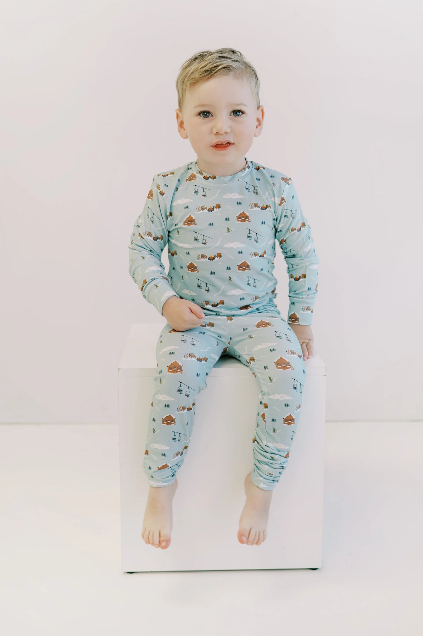 
                  
                    Child sitting on a white stool against a white background wearing a light blue bamboo lounge set with a ski resort themed print. 
                  
                